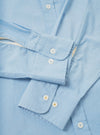 Close-up flat lay of the sky blue Daybrook shirt in chambray cotton, showing two buttoned barrel cuffs with rounded corners and white buttons with cross stitching. A cream garment tag is visible at the sleeve join. The placket with a single white button is visible at the top of the frame.
