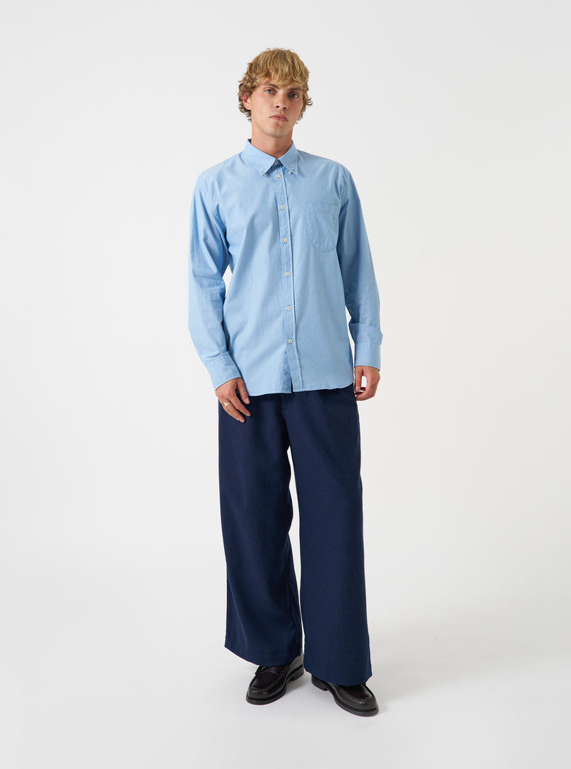 A male model poses against a plain light grey studio backdrop wearing the Sky Blue Prince Chambray Daybrook Shirt. The button-up cotton shirt features a spread collar, chest pocket, and long sleeves.