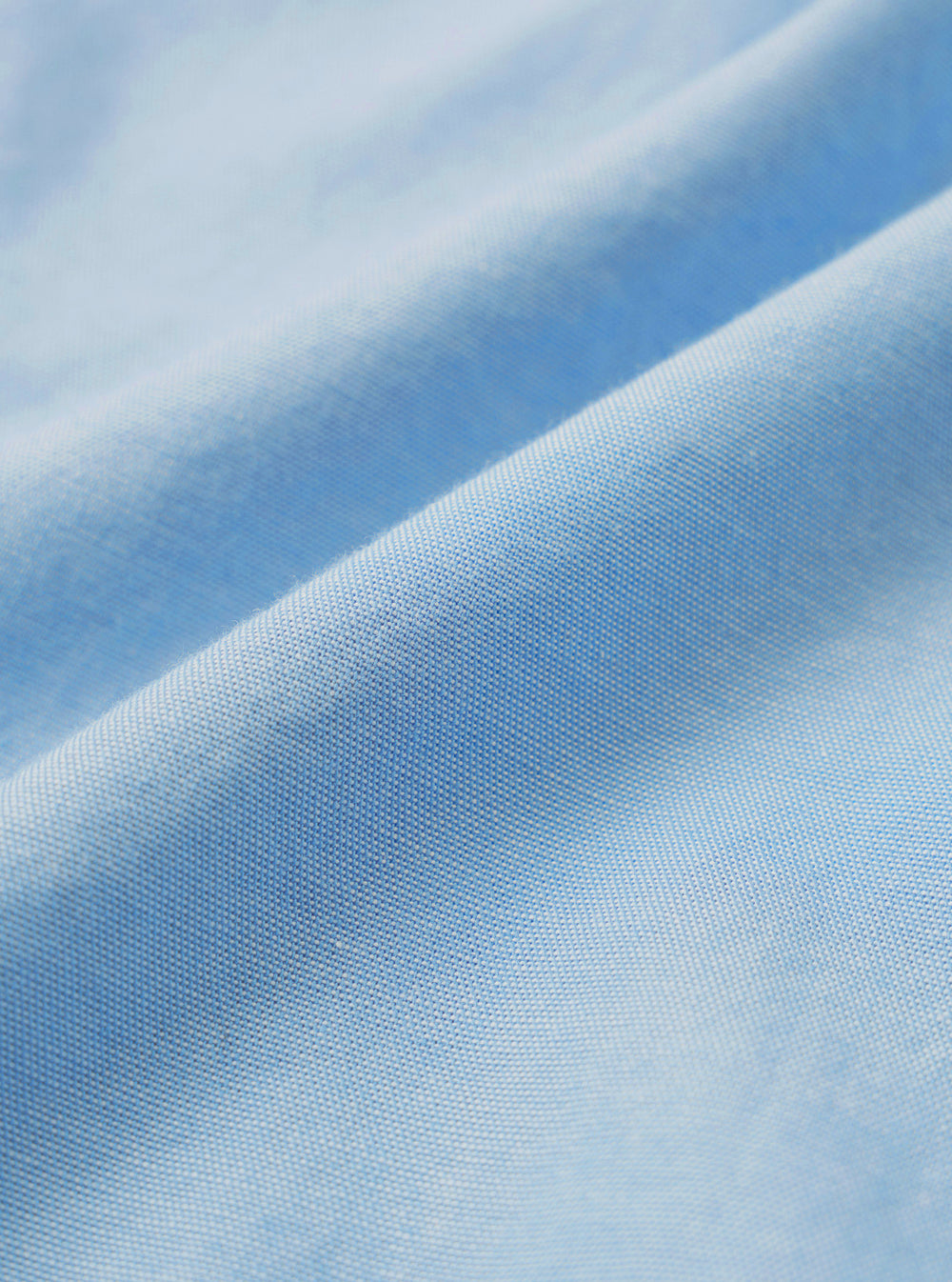 Close-up detail shot of the sky blue chambray fabric used in the Daybrook shirt. The tightly woven oxford-style weave is clearly visible, showing a fine two-tone texture with light and mid-blue threads. The fabric is softly draped, catching light across a diagonal fold.
