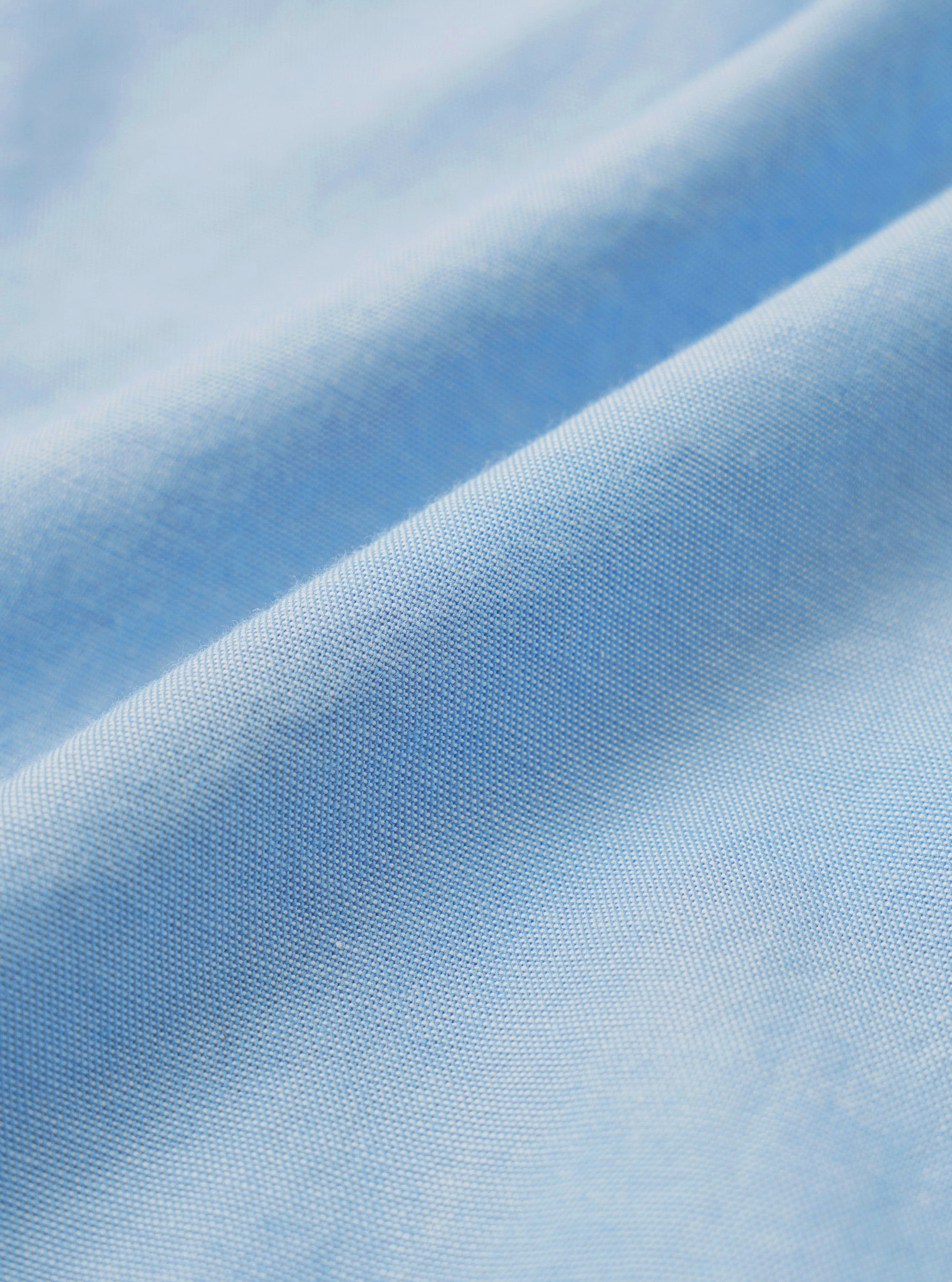 Close-up detail shot of the sky blue chambray fabric used in the Daybrook shirt. The tightly woven oxford-style weave is clearly visible, showing a fine two-tone texture with light and mid-blue threads. The fabric is softly draped, catching light across a diagonal fold.
