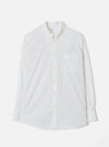 White Universal Works Daybrook shirt in prince chambray fabric, flat lay on a light grey backdrop. Button-down collar with a branded label at the neck, single patch chest pocket, full button placket with cream buttons, long sleeves with single-button cuffs, and a curved shirttail hem.