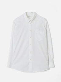 White Universal Works Daybrook shirt in prince chambray fabric, flat lay on a light grey backdrop. Button-down collar with a branded label at the neck, single patch chest pocket, full button placket with cream buttons, long sleeves with single-button cuffs, and a curved shirttail hem.