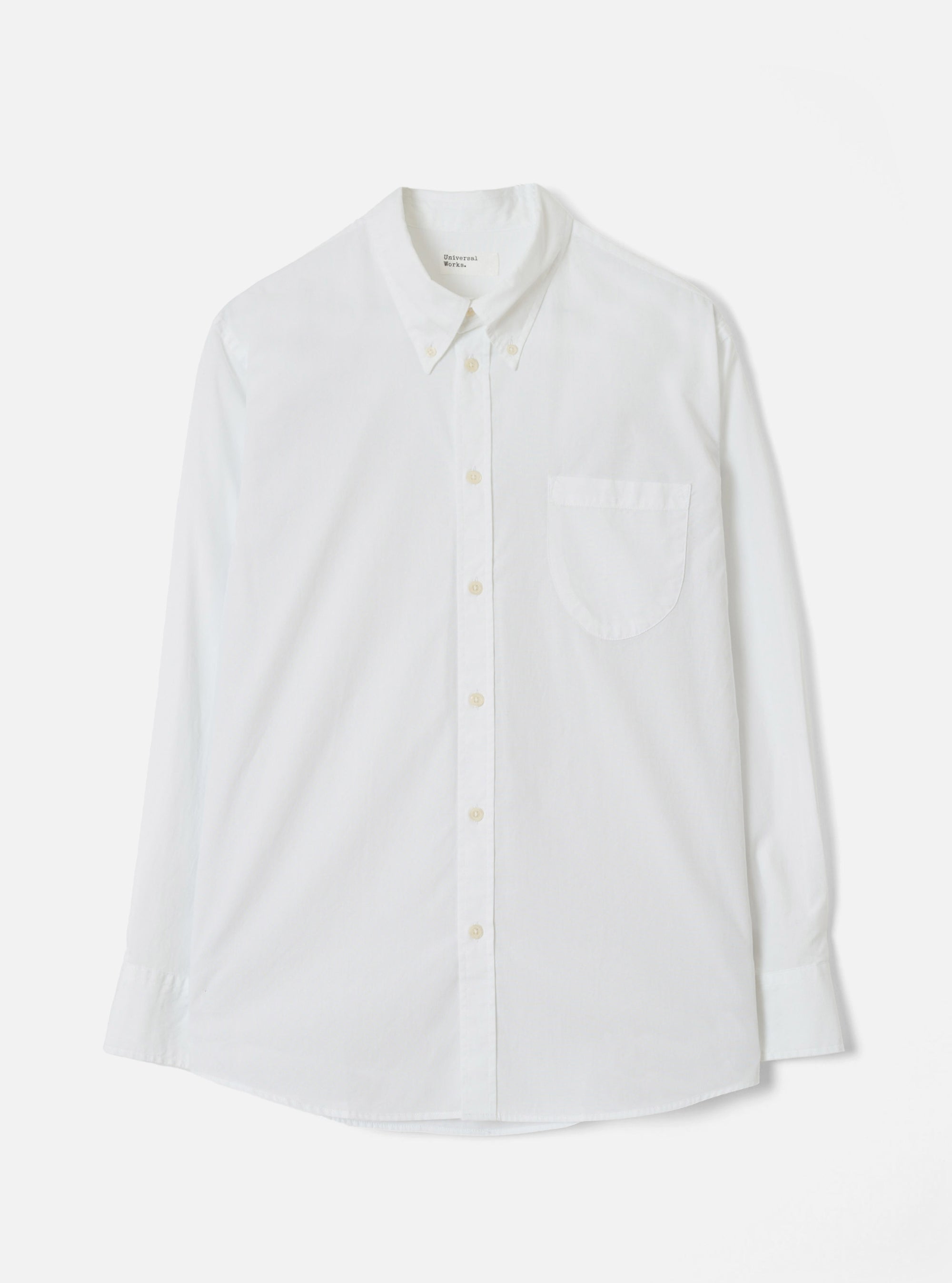 White Universal Works Daybrook shirt in prince chambray fabric, flat lay on a light grey backdrop. Button-down collar with a branded label at the neck, single patch chest pocket, full button placket with cream buttons, long sleeves with single-button cuffs, and a curved shirttail hem.