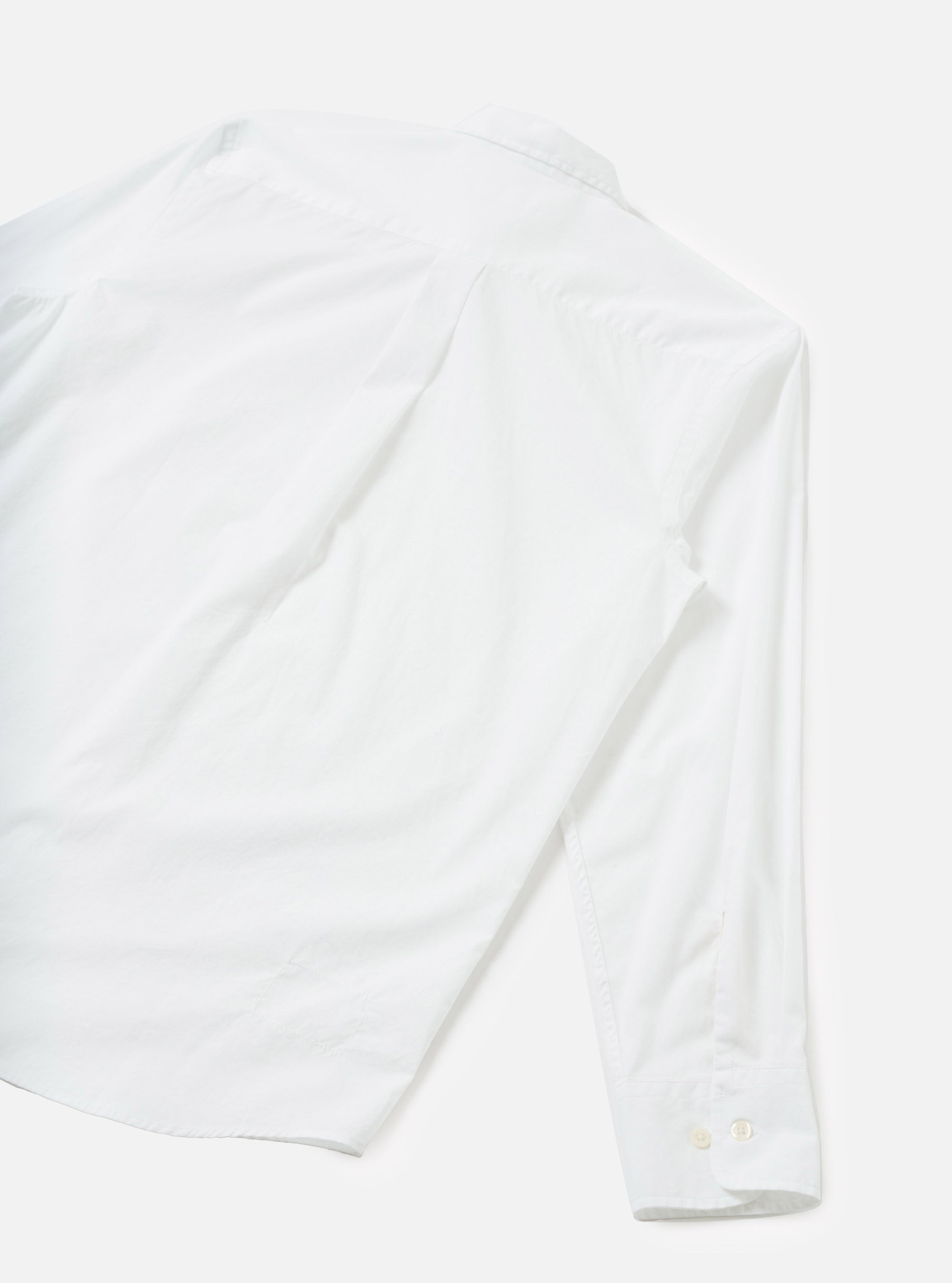 White chambray shirt laid flat on a light grey seamless surface, shot from above at an angle. The image shows the back of the shirt with one long sleeve extending to the lower right. A small collar is visible at the top. The buttoned cuff features two small white buttons. The fabric has a smooth, fine weave texture.