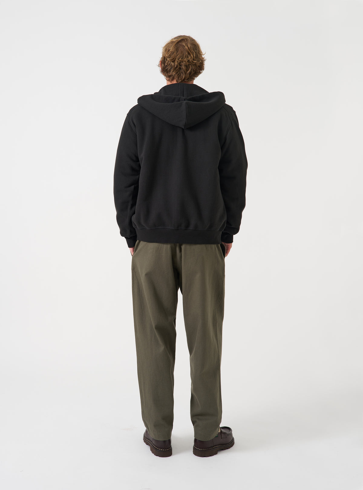 Side view of a model wearing the Black Brush Back Sweat Core Hoody against a neutral studio backdrop. The image shows the side profile of the black sweatshirt, demonstrating the cozy fleece construction, hood structure, and fit.