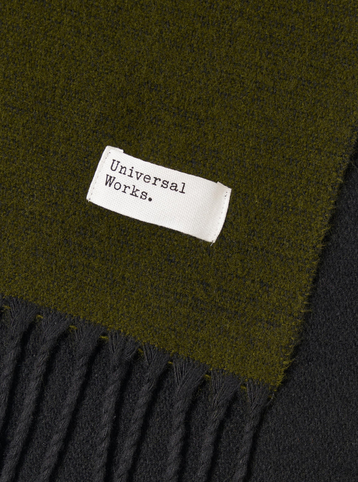 Detailed view of the Green/Black Double Sided Scarf draped flat against a plain studio backdrop. The shot showcases the contrast between the green and black sides, accentuating the soft texture and fringed trim along the edge.