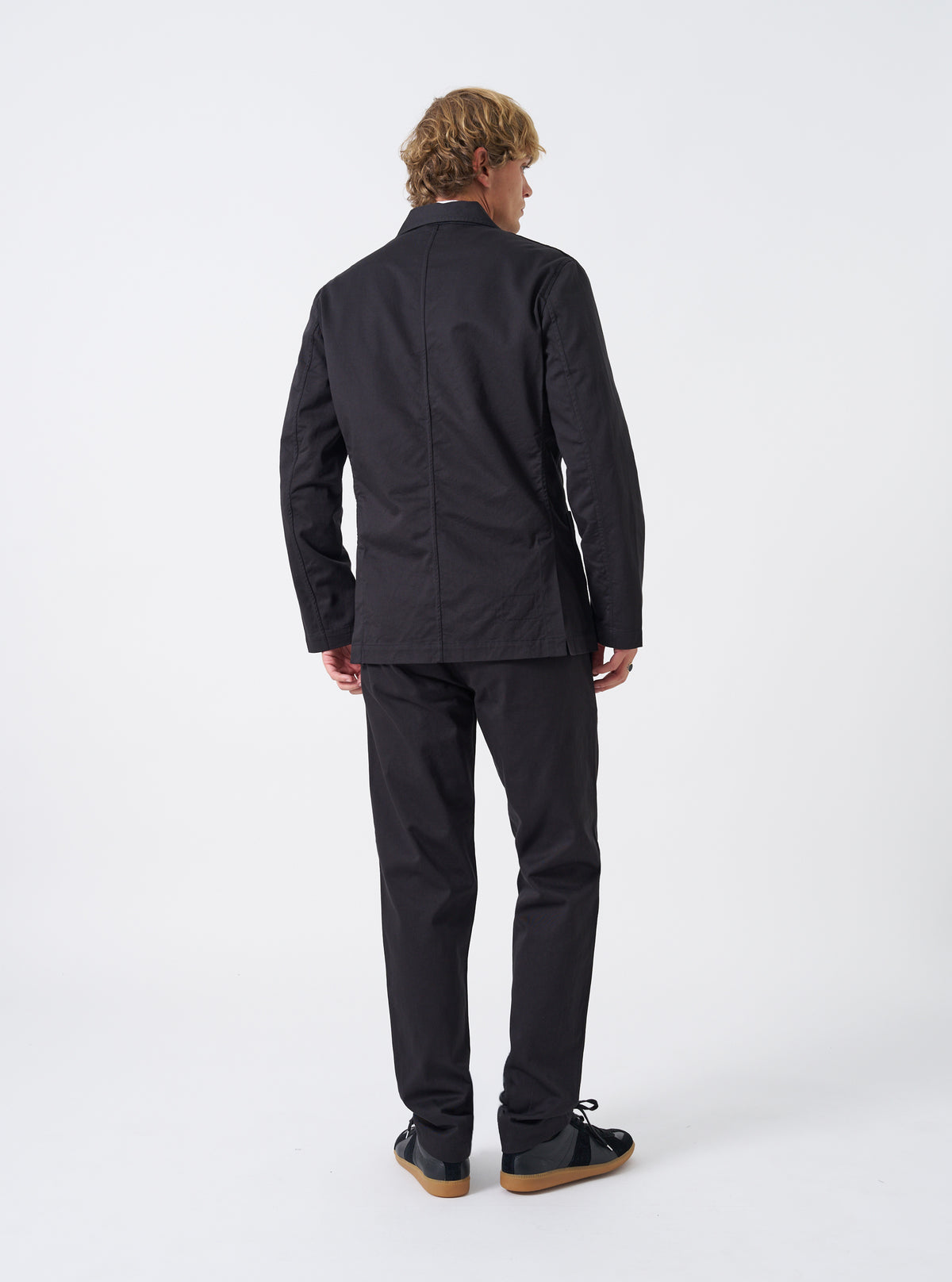 Side angled view of a male model wearing the Black Twill London Jacket in a studio setting with a clean neutral background, displaying the silhouette, sleeve length, and jacket drape when worn.