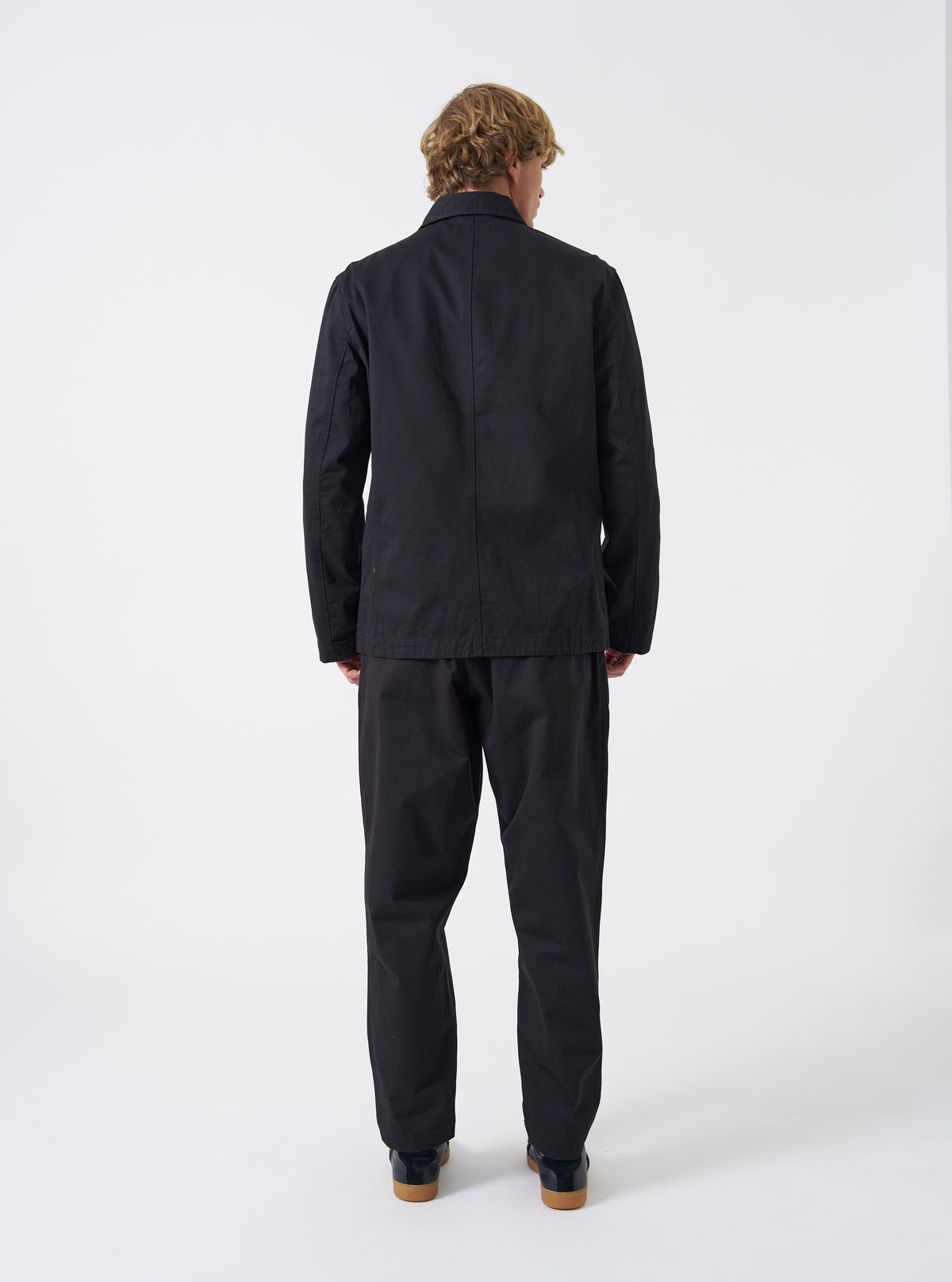 Side view of a male model wearing the Black Twill Bakers Chore Jacket against a neutral backdrop, demonstrating the profile silhouette, sleeve width, shoulder fit, and natural drape of the black twill coat.