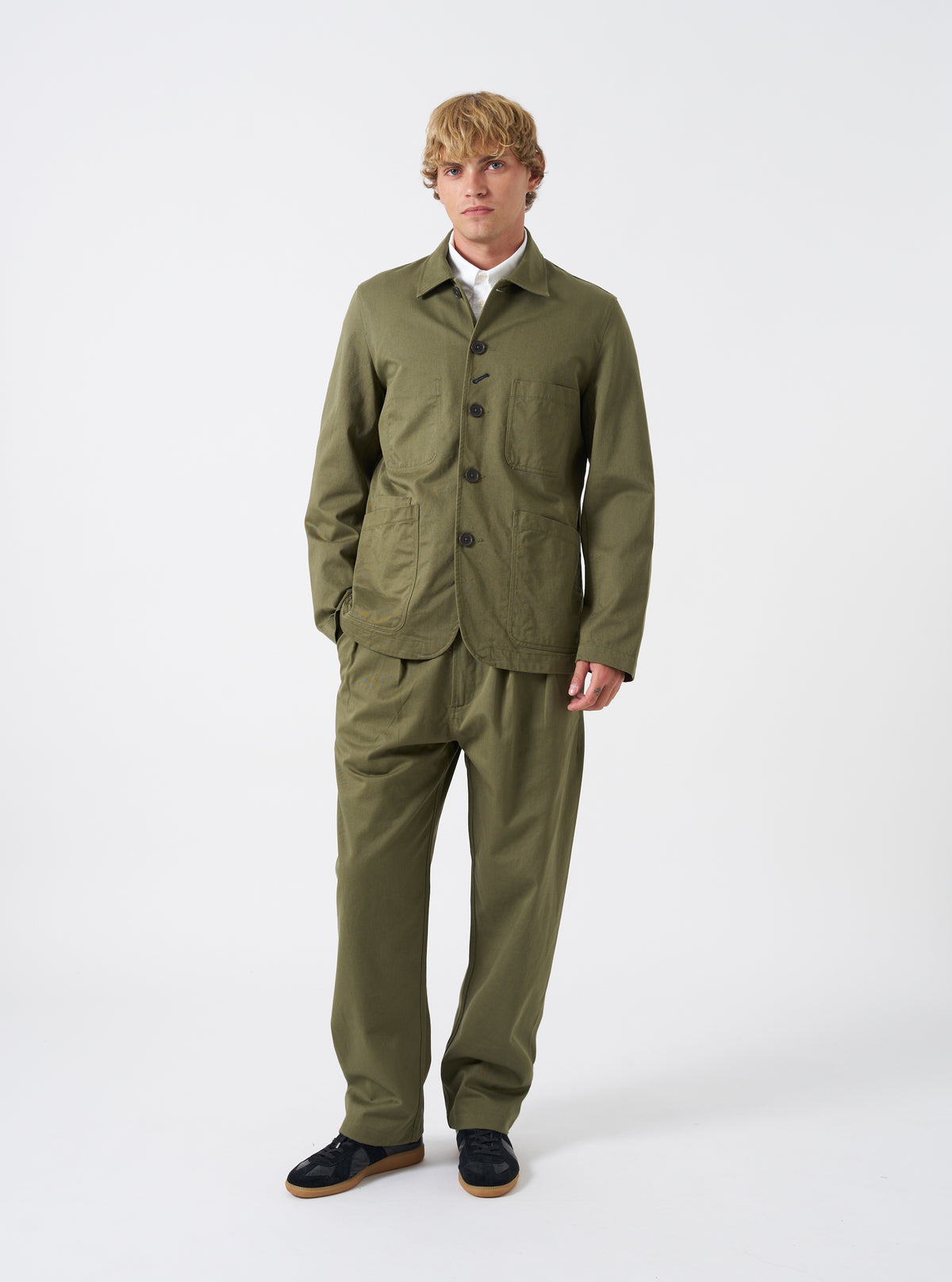 Front view of a model wearing the Light Olive Twill Bakers Chore Jacket in a bright studio setting. The model stands facing forward, showing the front button closure, collar, and utility pockets in light olive green.