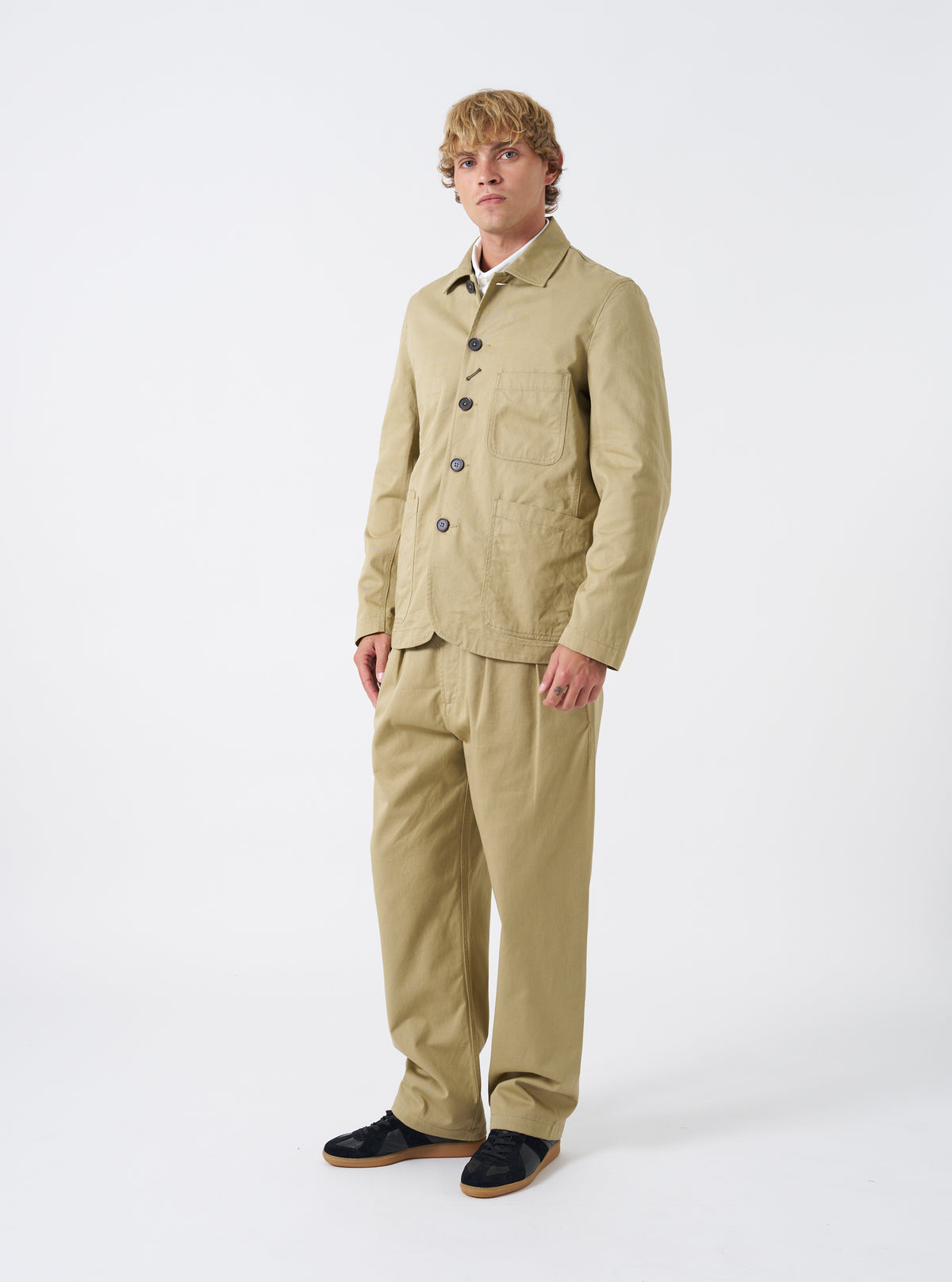 Front view of a model wearing the Sand Twill Bakers Chore Jacket in a plain studio setting. The model stands facing forward, showing how the sand tan jacket fits when buttoned over casual clothing.