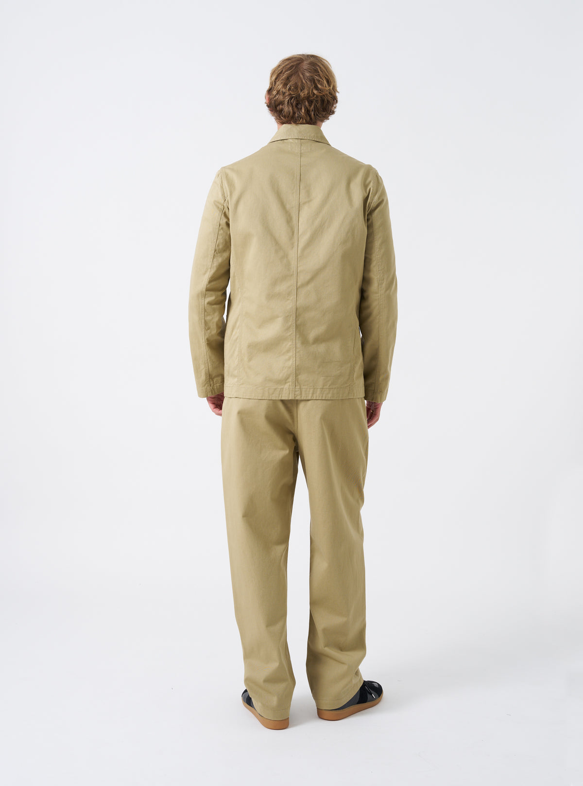 Rear view of a model wearing the Sand Twill Bakers Chore Jacket in a light studio background. The model stands facing away, displaying the drape, fit across the back, and overall silhouette.