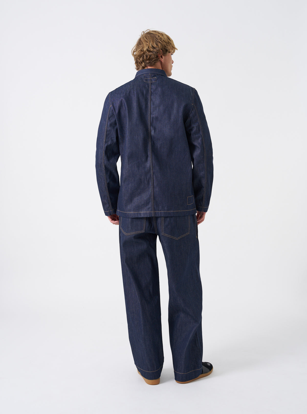 Back view of the Indigo 13oz Selvedge Denim Trucker Jacket on a plain background. The shot highlights the clean rear paneling, tailored shoulder seams, dark blue denim finish, and adjustable waist tabs located near the bottom hem.