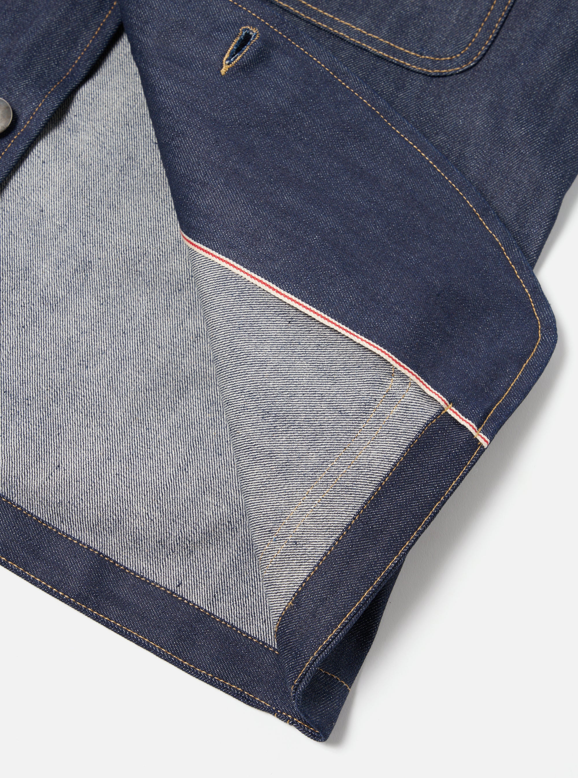 Close-up of the Indigo 13oz Selvedge Denim Bakers Jacket on a white surface. Shows the denim texture, red selvedge detail, and tan stitching. 
