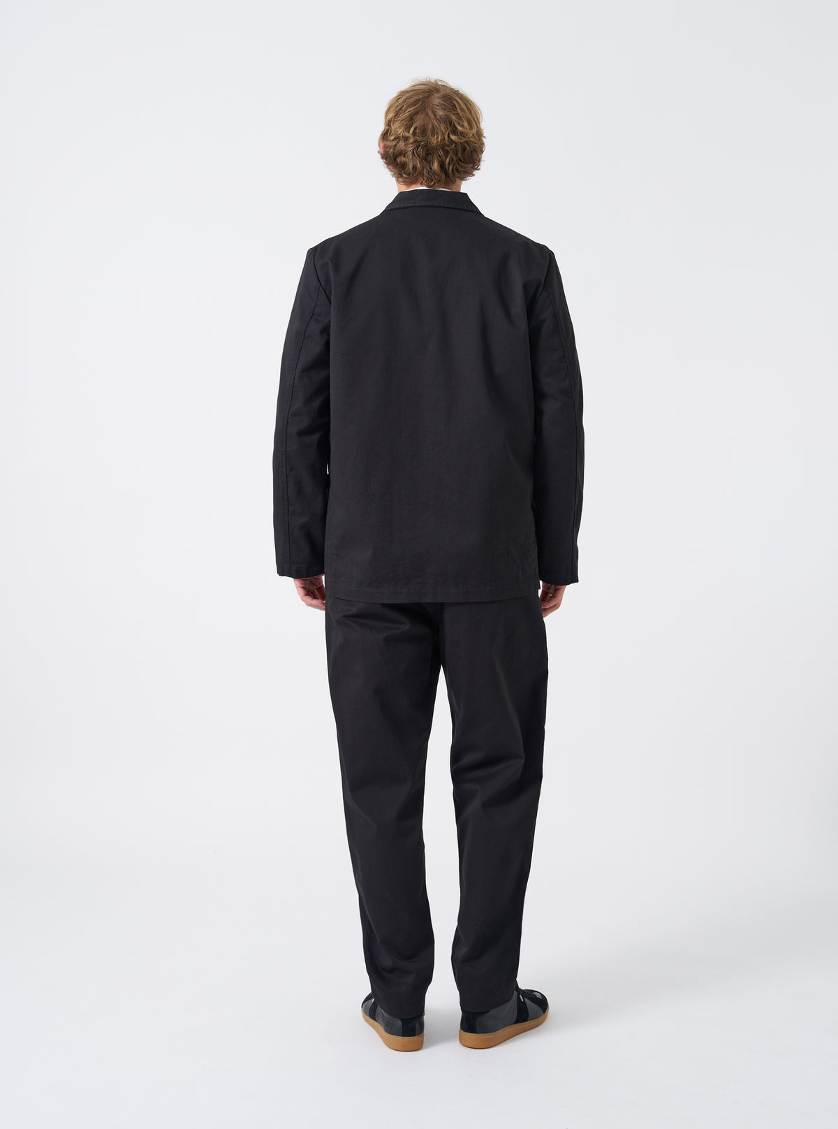 A male model wearing the Black Twill Three Button Jacket in an unbuttoned pose against a plain studio background, demonstrating how the jacket drapes over trousers and a layered shirt.