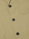 Close-up of the sand twill three button jacket on a flat surface. The jacket is a light tan color with dark brown buttons. The background is a plain white.
