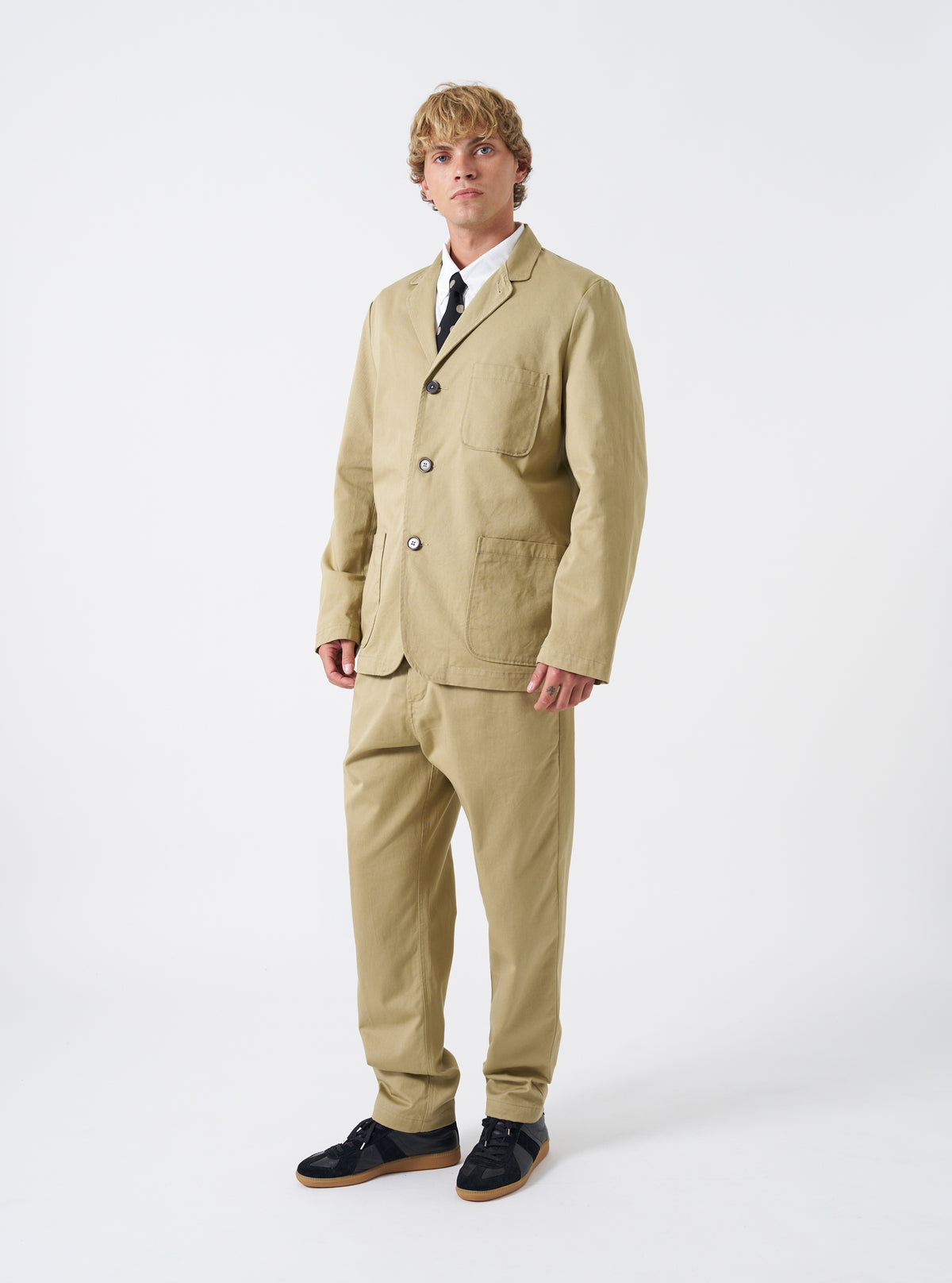 A male model is wearing the Sand Twill Three Button Jacket in a full-length studio shot against a plain white background. The jacket is styled open over a neutral shirt with matching trousers.