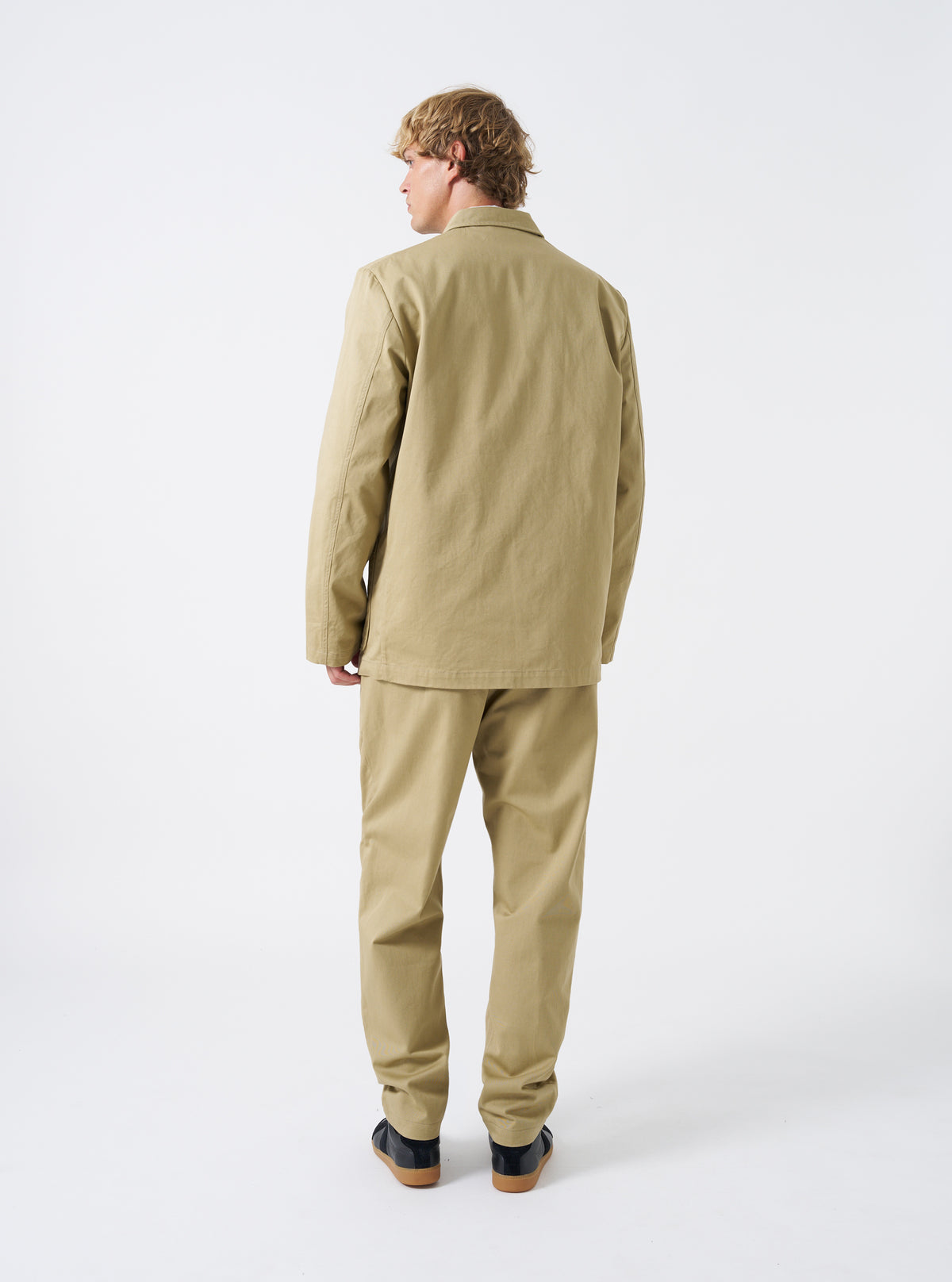 Side profile view of a male model wearing the Sand Twill Three Button Jacket against a clean studio background. Demonstrates the natural shoulder line, tailored sleeve cut, and relaxed silhouette.