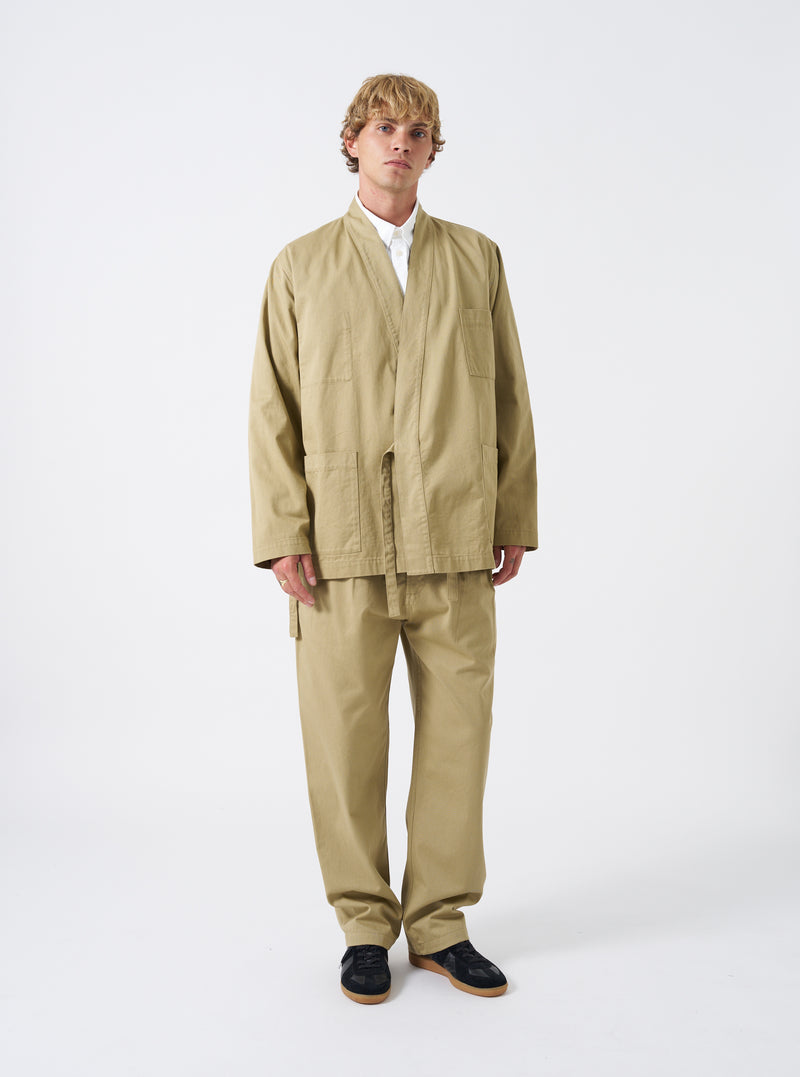 A male model is wearing the Sand Twill Kyoto Work Jacket in a studio setting against a light background. He wears the jacket closed with side fabric ties paired with casual trousers.