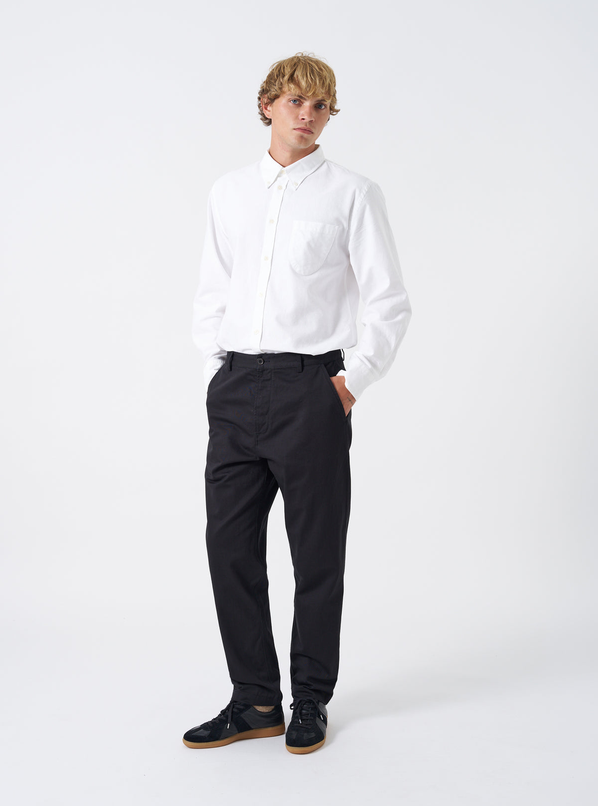 Full front shot of a model wearing the Black Twill Military Chino on a clean white background. The model wears the black trousers paired with casual shoes, demonstrating the comfortable mid-rise fit and neat taper toward the ankles.