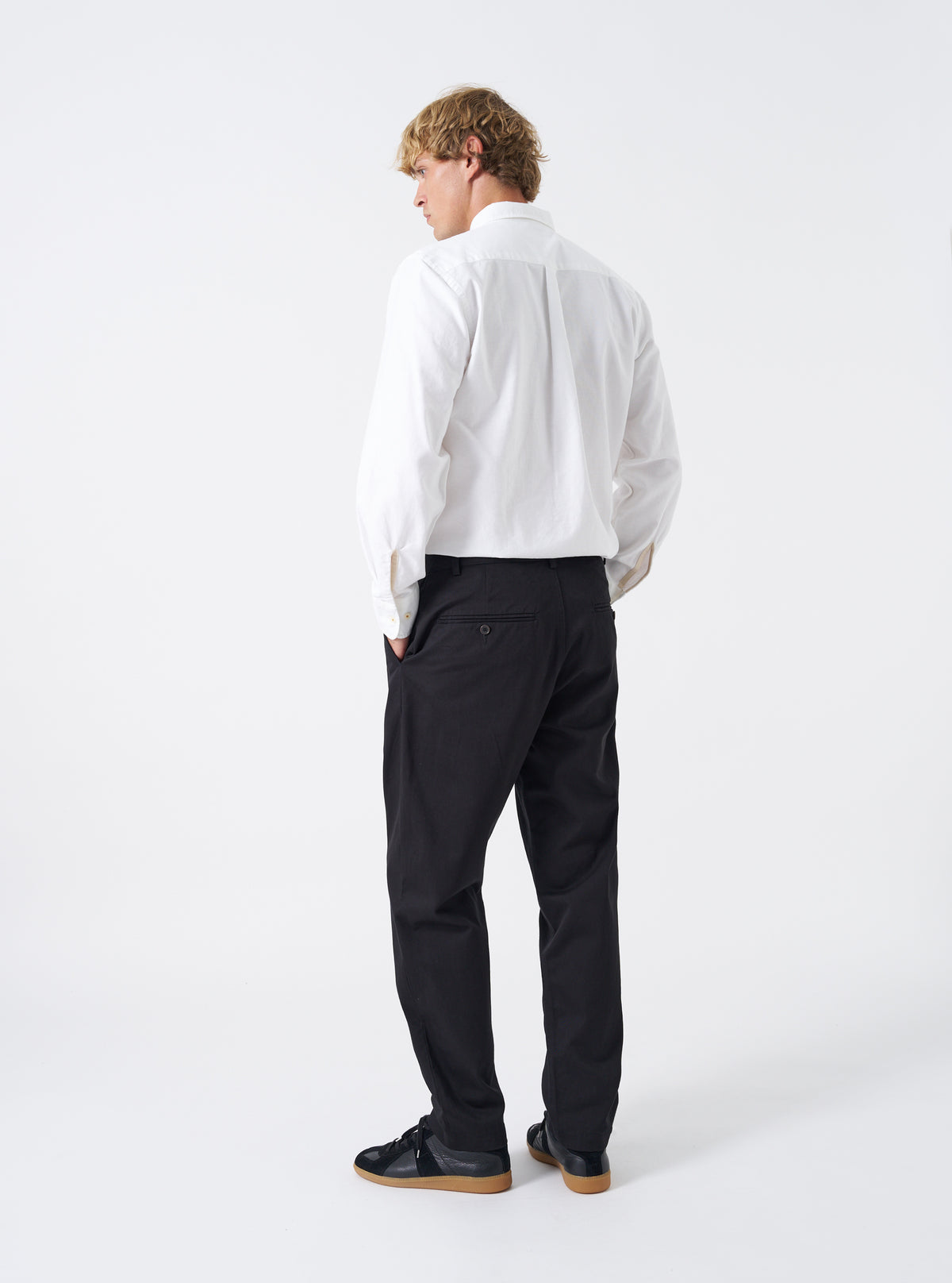 Rear view of a model wearing the Black Twill Military Chino against a plain white studio backdrop. Highlights how the dark cotton twill trousers fit through the seat and thighs, featuring rear jetted button pockets and clean hem finish.