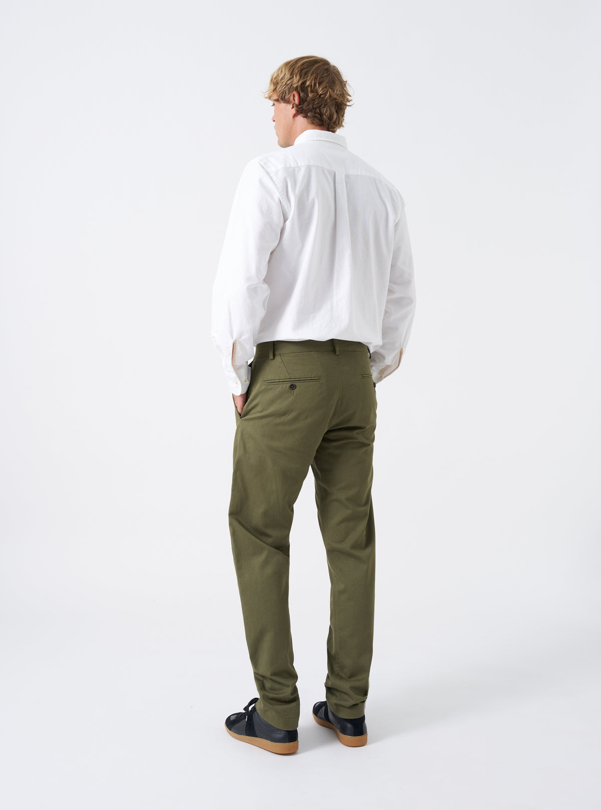 Rear shot of a model wearing the Light Olive Twill Military Chino against a white background. Shows the back fit of the light olive cotton trousers, highlighting the rear buttoned flapless pockets and tapered fit down to the ankles.