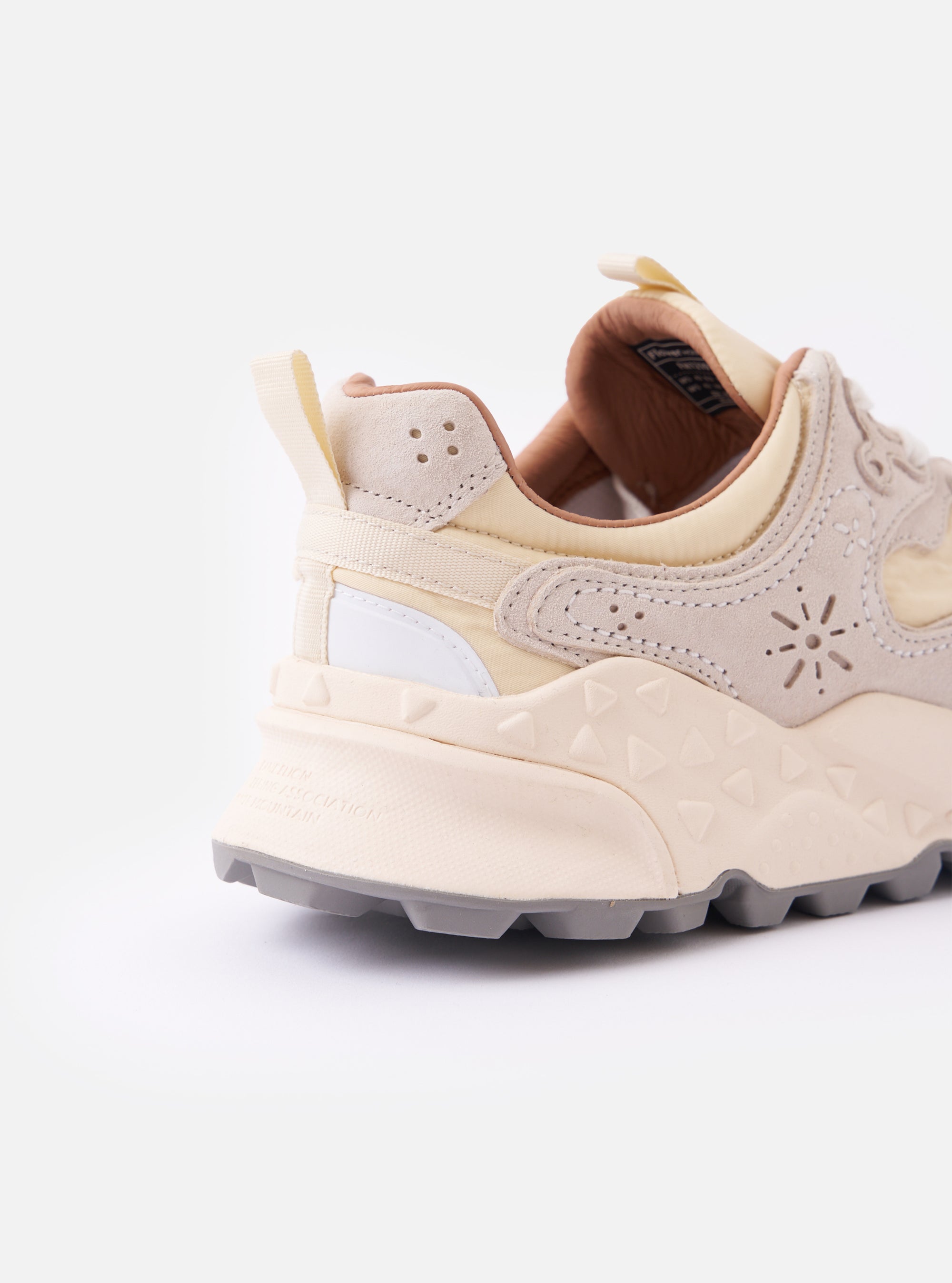 The Flower Mountain Milk/Light Beige Suede/Nylon Rulis Uni sneaker is shown from a side profile against a plain white background. The shoe features a combination of milk-colored technical nylon and light beige suede overlays with decorative floral-shaped perforations and zigzag stitching. It has a chunky, off-white molded rubber sole with a textured tread, white cotton laces, and a pull tab at the heel.