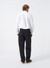 Rear view shot of a model wearing the Black Twill Curved Pant on a bright neutral backdrop. The model stands wearing the black trousers, highlighting the back pockets, waist fitting, and tapered leg opening at the ankles.
