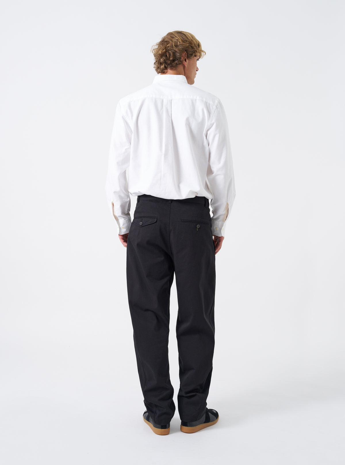 Rear view shot of a model wearing the Black Twill Curved Pant on a bright neutral backdrop. The model stands wearing the black trousers, highlighting the back pockets, waist fitting, and tapered leg opening at the ankles.
