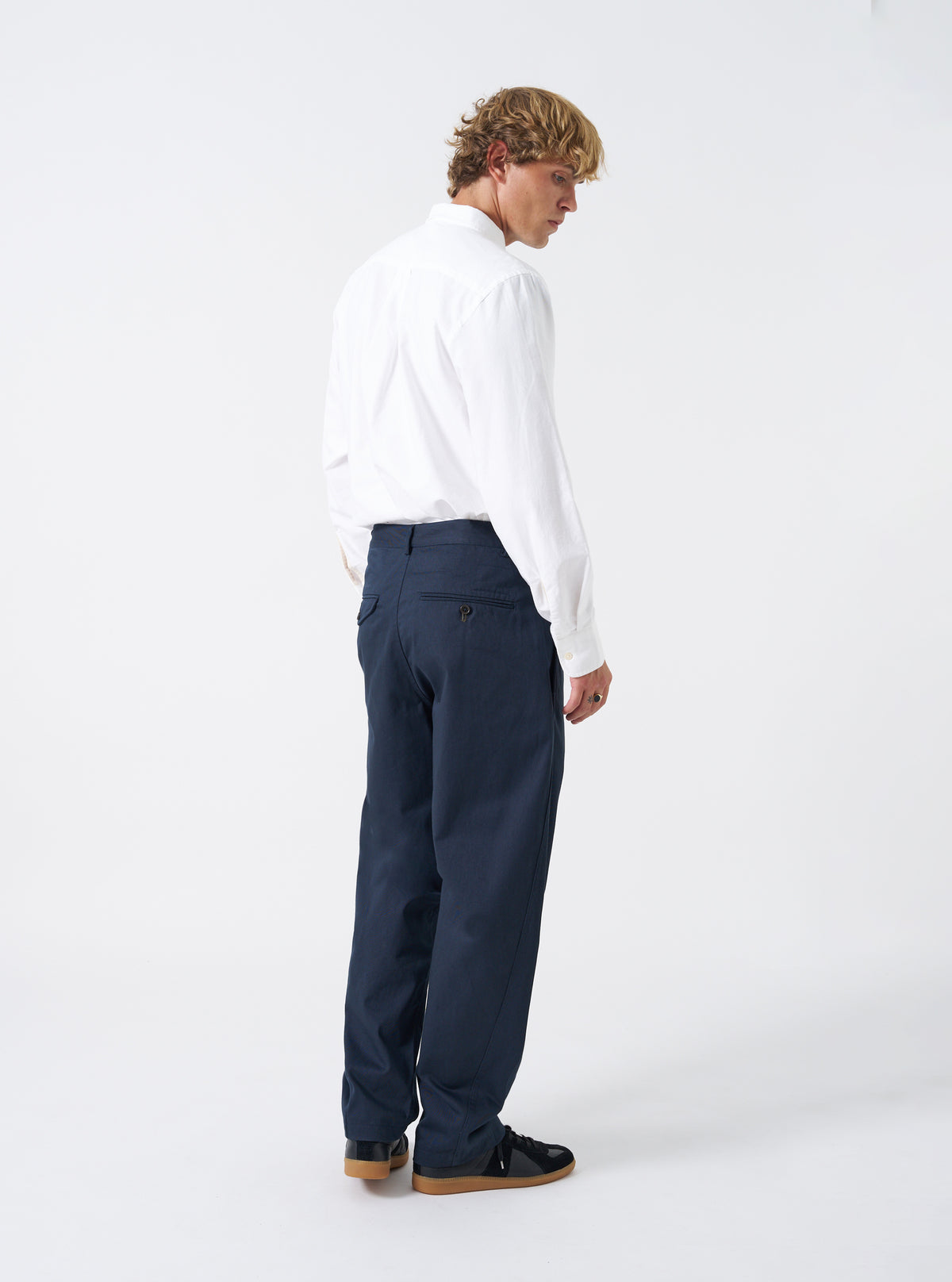 Rear view of a model wearing the Navy Twill Curved Pant against a plain white studio backdrop. The model wears the navy trousers, emphasizing the rear jetted and flap pockets alongside the signature curved silhouette.