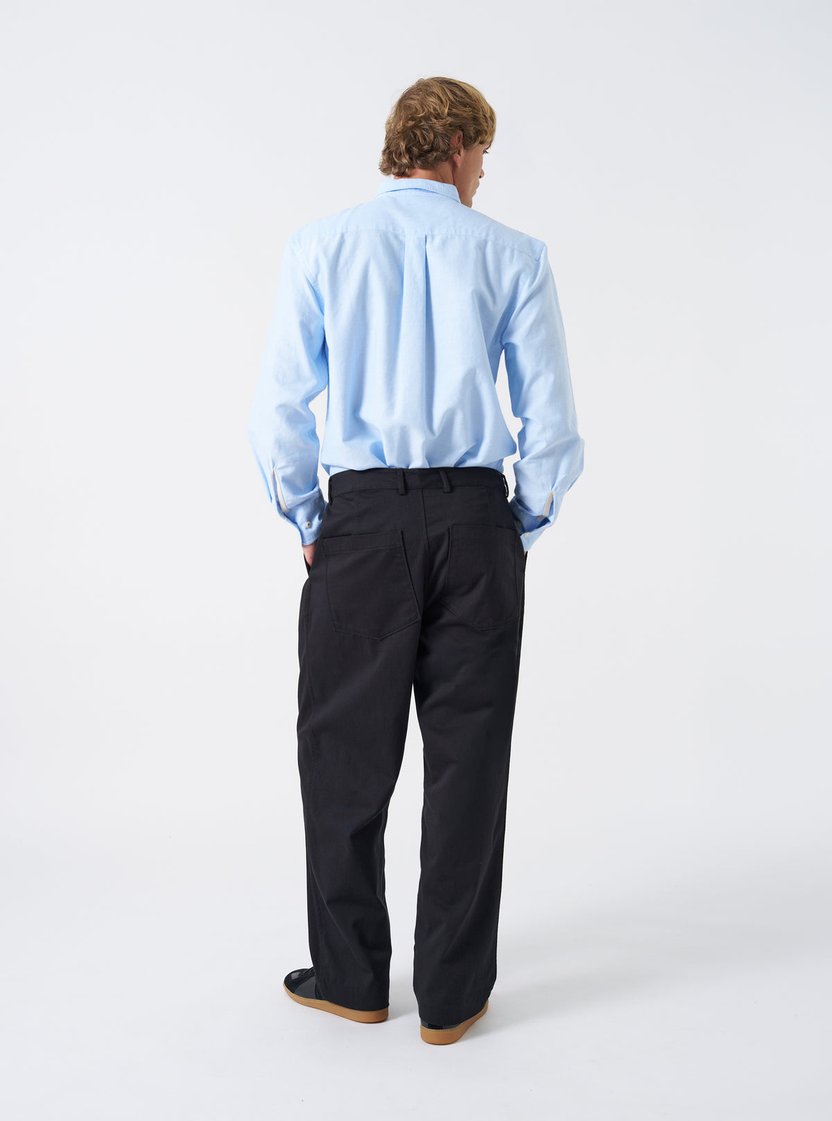 Back view shot of a model wearing the Navy Twill Duke Pant against a clean neutral studio backdrop. The model stands wearing the dark navy pants, demonstrating the relaxed drape, back patch pockets, and slight leg taper.