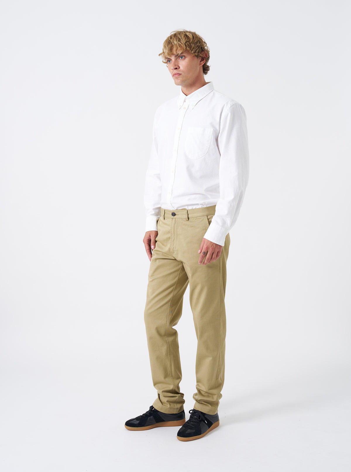Front view of a male model wearing the Sand Twill Aston Pant standing against a neutral studio backdrop. Shows the low-rise waist, slim straight-leg fit, side pockets, and styled appearance of the sand-colored twill trousers.