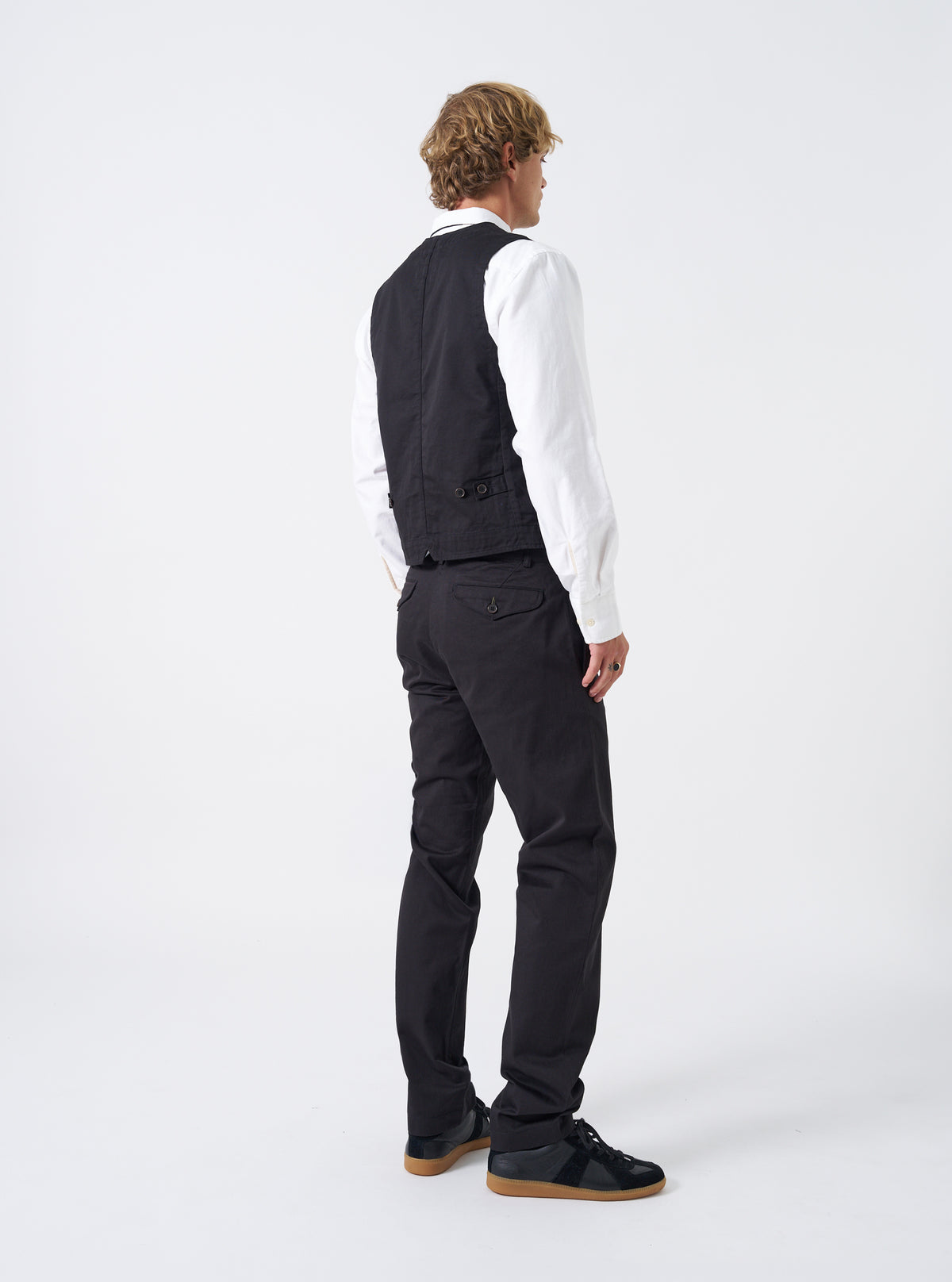 Rear perspective of a model wearing the Black Twill Aston Pant on a clean off-white backdrop. The model is shown standing in the slim black trousers, emphasizing the tailored fit through the legs and rear pocketing.