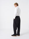 Side angle shot of a model wearing the Black Twill Double Pleat Pant on a neutral background. Demonstrates the relaxed silhouette, room through the leg, and precise waistband tailoring on the body.