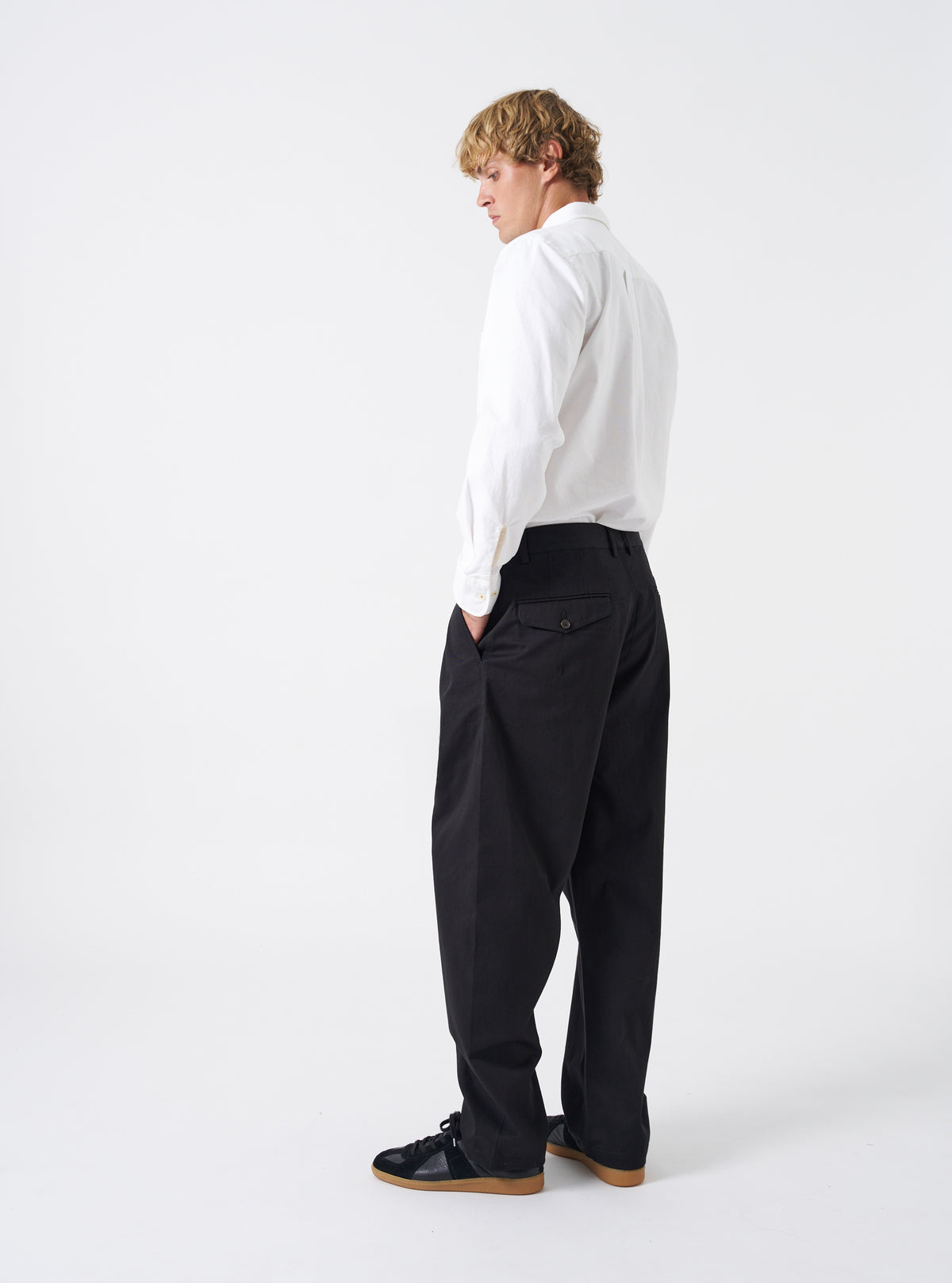 Side angle shot of a model wearing the Black Twill Double Pleat Pant on a neutral background. Demonstrates the relaxed silhouette, room through the leg, and precise waistband tailoring on the body.