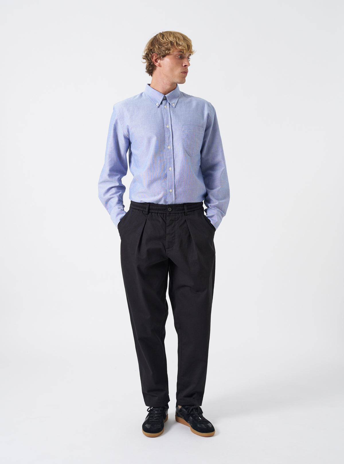 Full-length view of a model wearing the Black Twill Pleated Track Pant against a plain backdrop. Displays the tapered leg silhouette, relaxed fit around the waist, and drawstring closure styled with shoes.