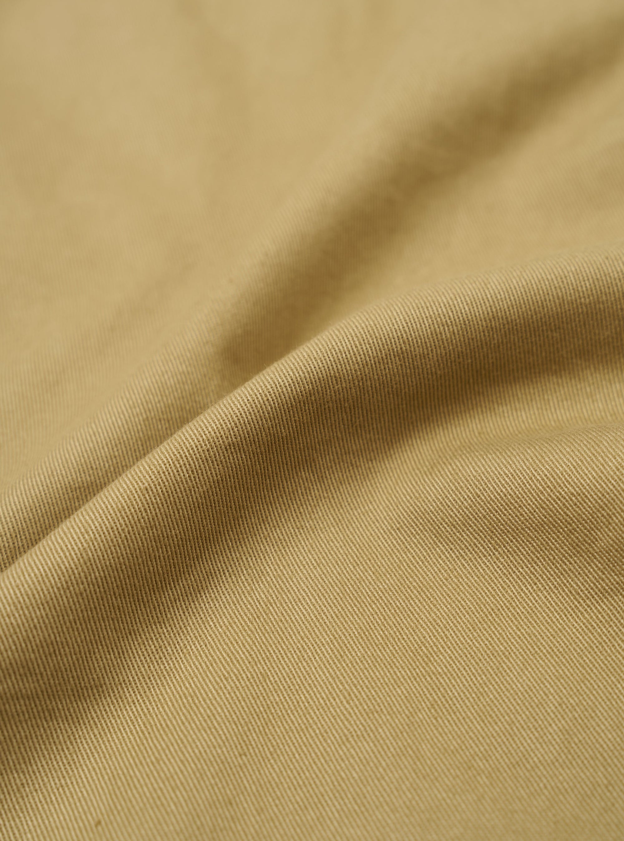 A close-up swatch of the Sand Twill Beach Short highlights the texture and diagonal weave of the sand-colored twill fabric. The material fills the frame, providing a detailed view of the garment's color and construction.