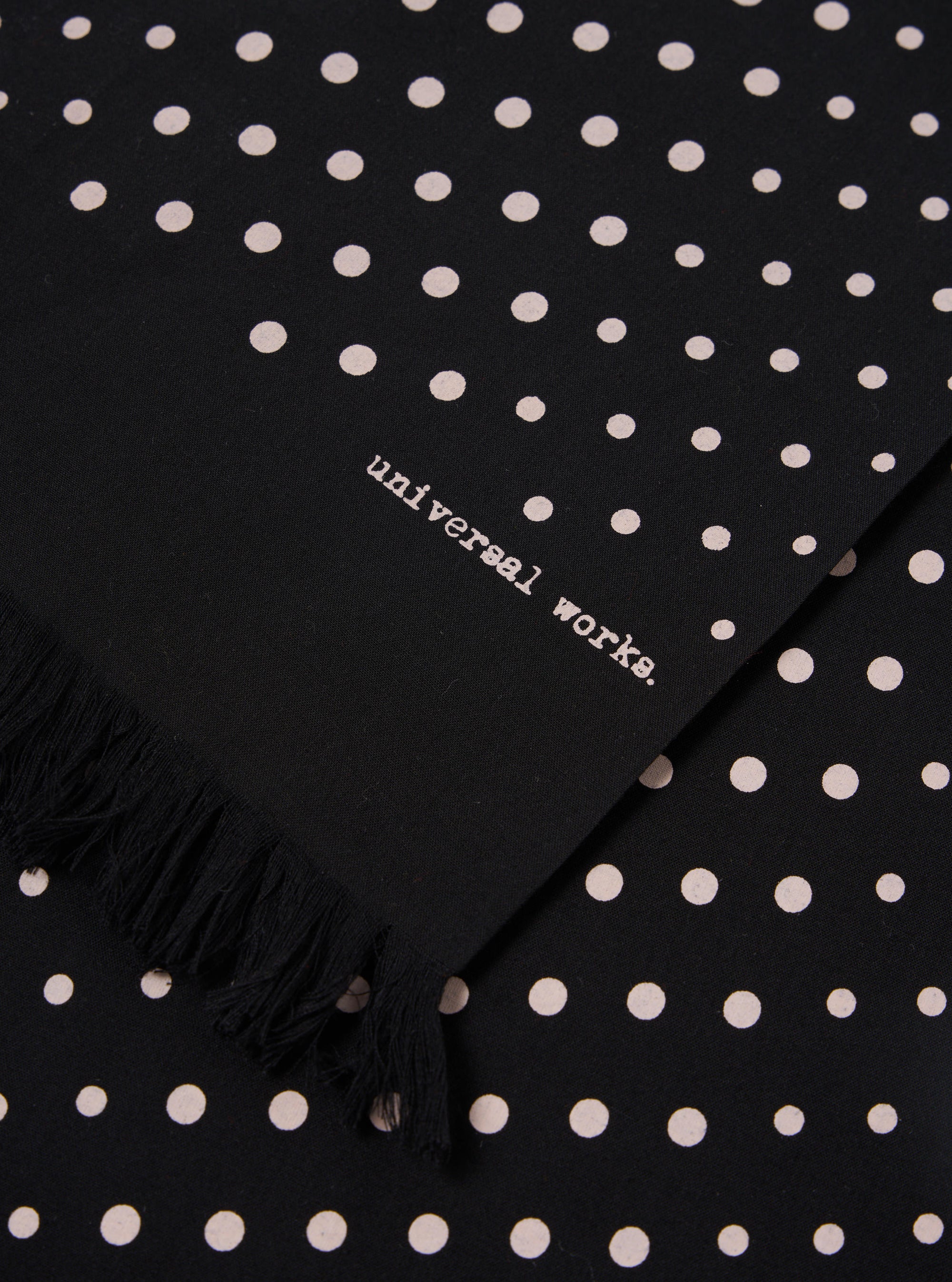 Close-up of a Black Dot Print Short Scarf with fringe. The scarf has white dots and the words "universal works."
