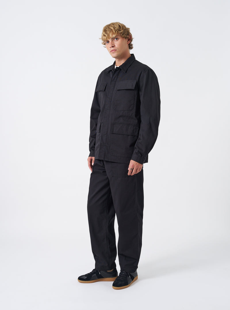 A model wears the Black Twill MW Fatigue Jacket in a studio setting against a plain background. The full-length shot shows the jacket buttoned up, highlighting its relaxed military fit and four front flap patch pockets.