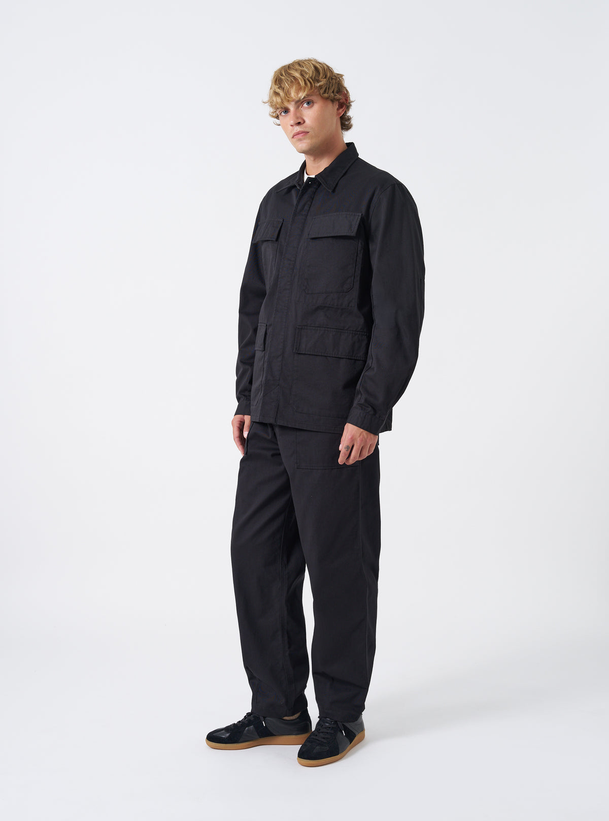 A model wears the Black Twill MW Fatigue Jacket in a studio setting against a plain background. The full-length shot shows the jacket buttoned up, highlighting its relaxed military fit and four front flap patch pockets.