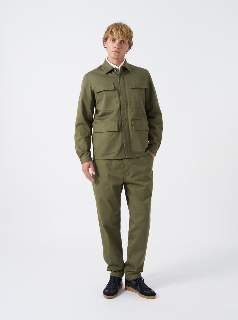 A model stands against a plain studio background wearing the Light Olive Twill MW Fatigue Jacket. The front-facing image showcases the relaxed M65 military silhouette, four utility pockets, and full buttoned placket.