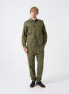 A model stands against a plain studio background wearing the Light Olive Twill MW Fatigue Jacket. The front-facing image showcases the relaxed M65 military silhouette, four utility pockets, and full buttoned placket.
