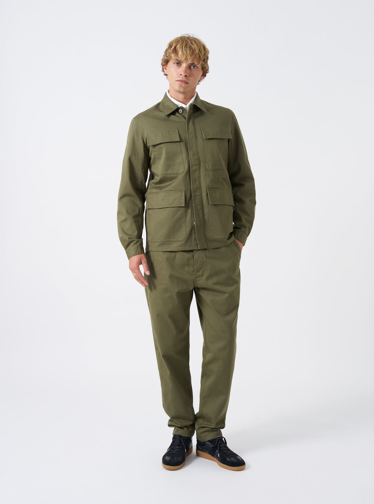A model stands against a plain studio background wearing the Light Olive Twill MW Fatigue Jacket. The front-facing image showcases the relaxed M65 military silhouette, four utility pockets, and full buttoned placket.