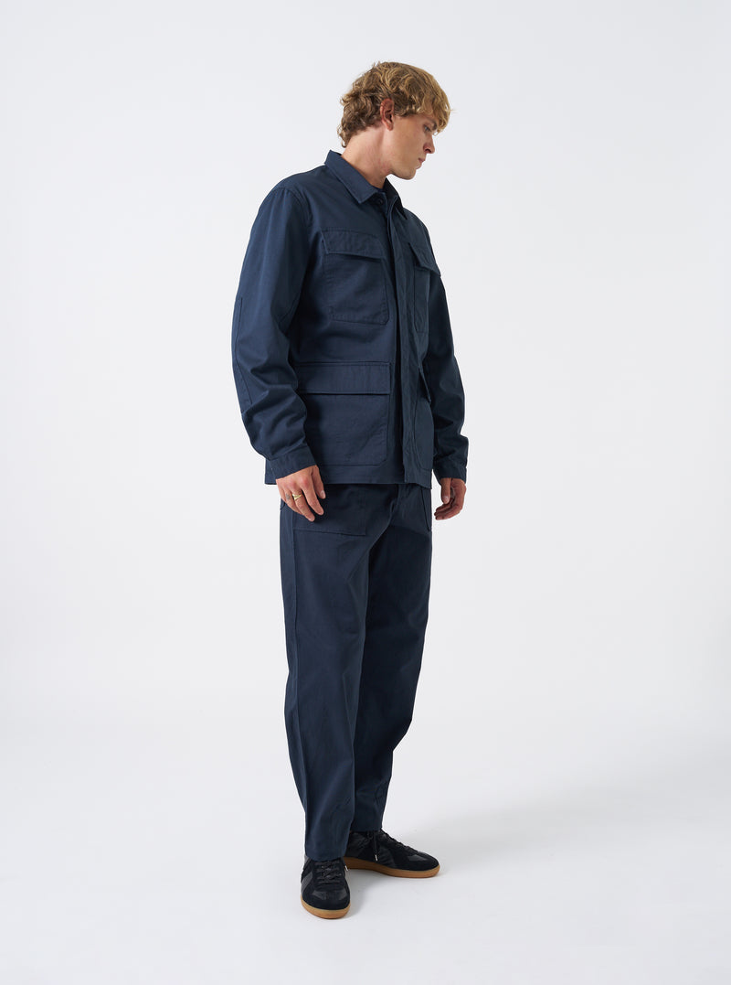 A model wearing the Navy Twill MW Fatigue Jacket stands in a studio setting against a plain background. The front view highlights the dark blue cotton twill fabric, concealed button front, and four front flap patch pockets.