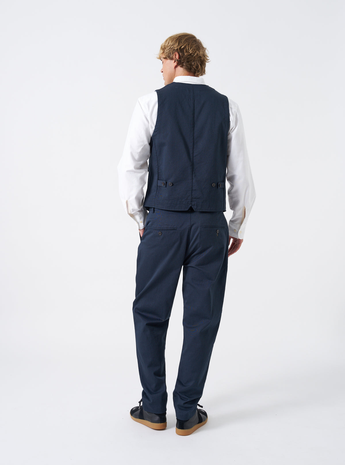 A model stands facing sideways against a neutral studio backdrop wearing the Navy Twill Field Waistcoat. The image displays the side profile, armhole shape, layered fit, deep navy twill material, and functional front pocket placement clearly.