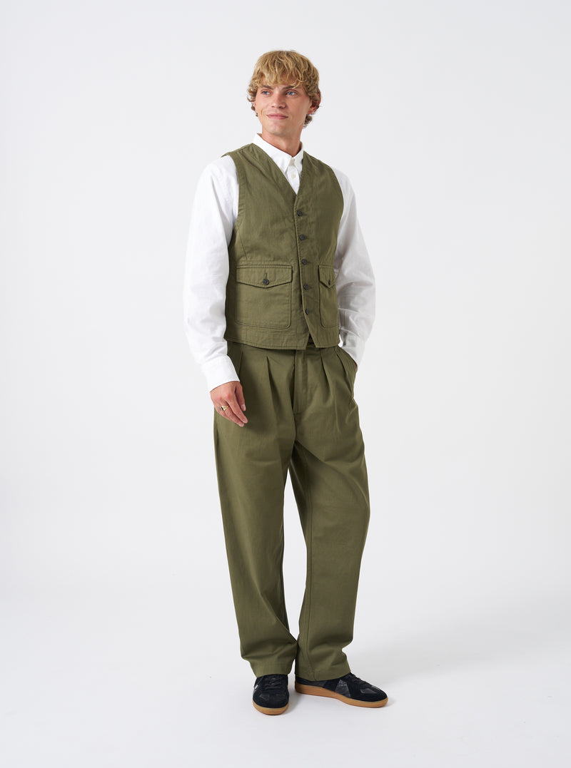 A model stands against a plain studio backdrop wearing the Light Olive Twill Field Waistcoat over a collared shirt. The shot displays the relaxed fit, front button closure, distinct olive twill fabric shade, and front utility patch pockets.