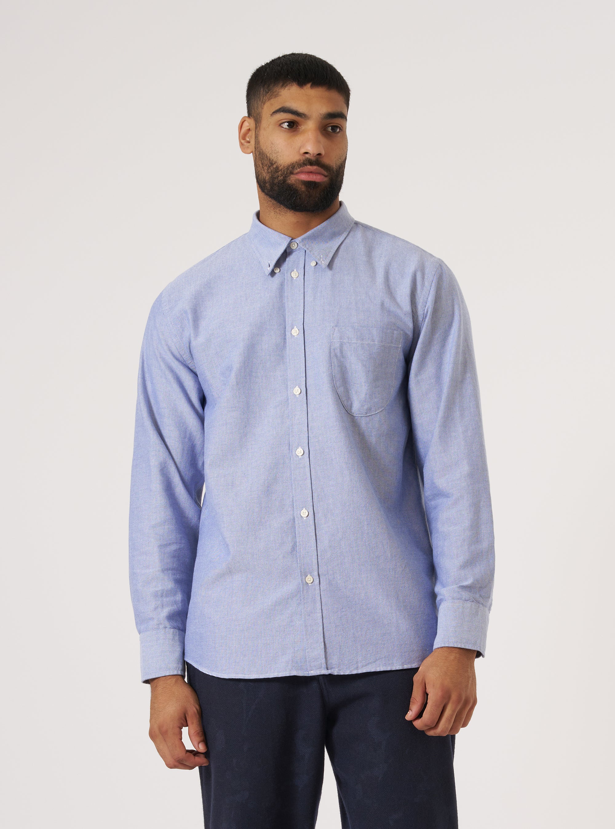 A man stands against a white backdrop, modeling the Blue Oxford Daybrook Shirt. The shirt is light blue, buttoned, and has a pocket. He wears dark pants.