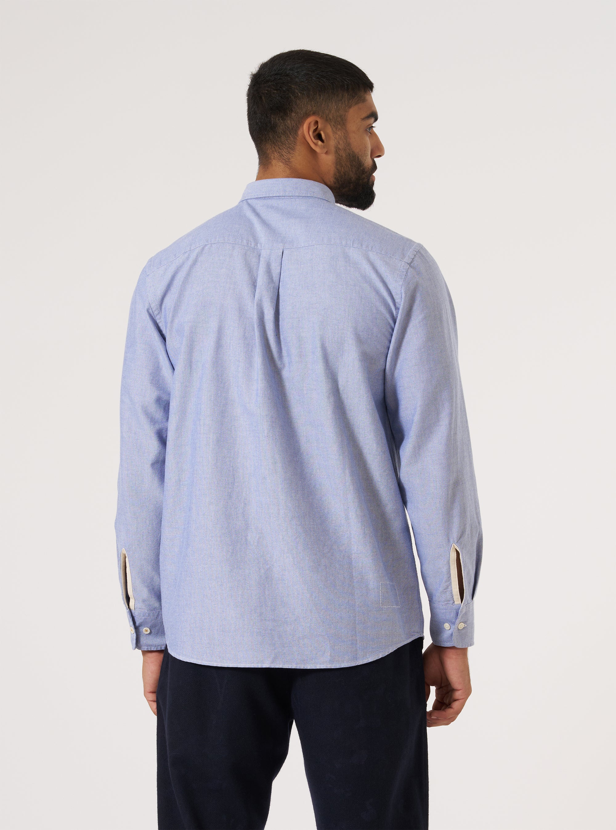 A man stands against a white backdrop, showcasing the back of a Blue Oxford Daybrook Shirt. The shirt is paired with dark trousers. 