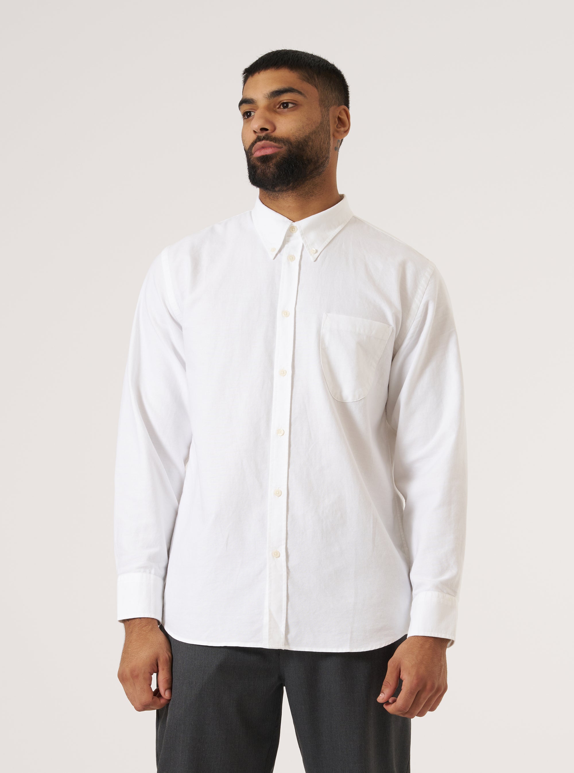 A man poses in front of a white background wearing a White Oxford Daybrook Shirt. The shirt is buttoned up and he is also wearing grey pants.