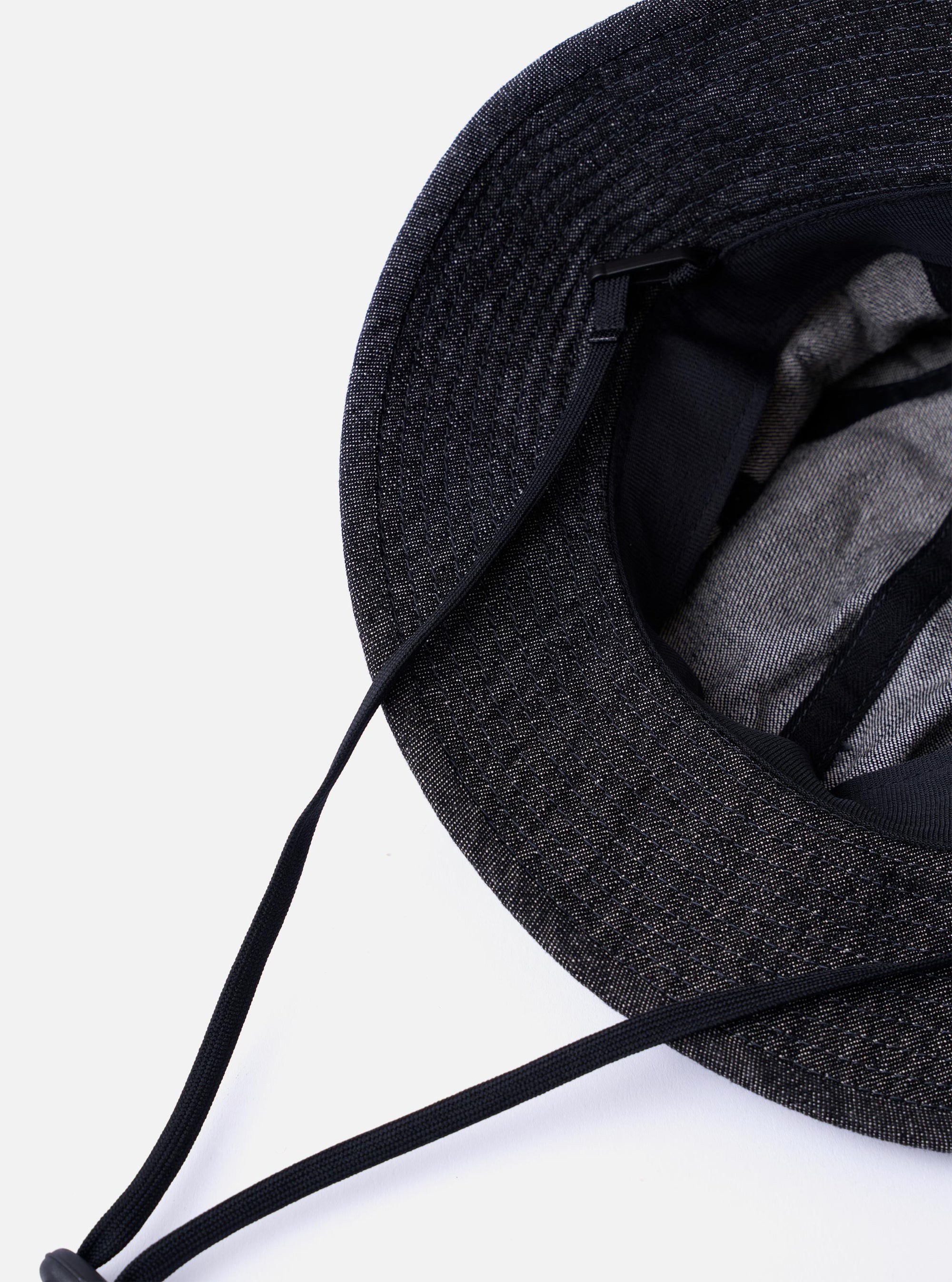 The cableami Black 10oz Denim Metro Hat is shown from a side angle as a flat lay against a plain white background. The image highlights the hat's rounded crown construction and the downward-sloping brim made from black denim with a visible natural slub texture. A black adjustable cord with a toggle is attached to the hat.