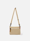 The master-piece x Universal Works Beige Canvas Sacoche is shown as a flat lay against a white background. The small rectangular shoulder bag is crafted from a durable beige canvas fabric and features a dual-branded logo patch on the front, a black adjustable shoulder strap with metal hardware, and a main compartment with a secure closure.